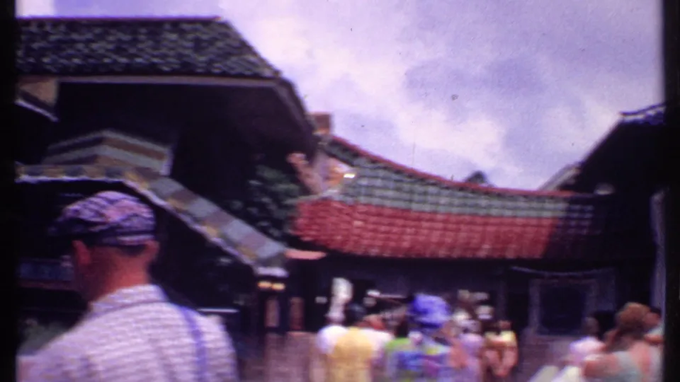 1969 Bahamas frame showing tourists in period clothing walking through a courtyard beside tiled-roof attraction buildings.
