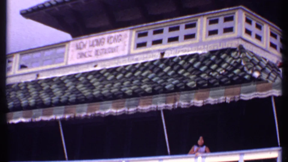 1969 Bahamas frame of the New Hong Kong Chinese Restaurant exterior, with tiled roof, awning, balcony railing, and a woman above.