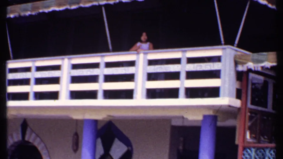 1969 Bahamas home-movie view of the restaurant balcony, awning, purple columns, arched doorway details, and a woman leaning at the rail.