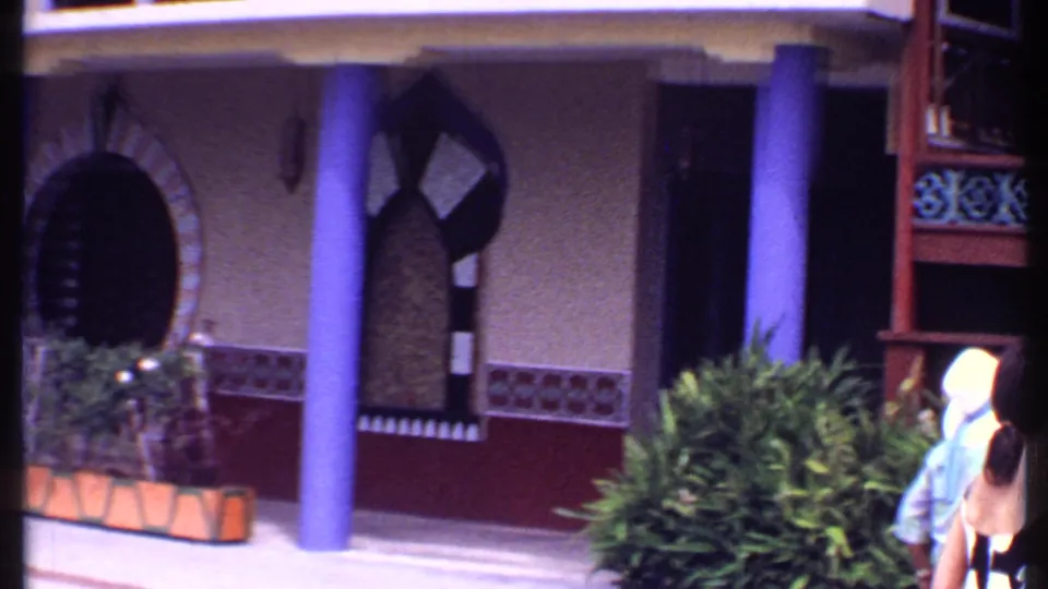 1969 Bahamas frame showing the restaurant facade with purple columns, arched doorways, decorative trim, greenery, and tourists at the right edge.