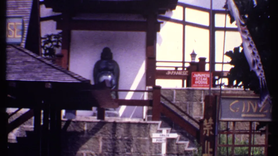 1969 Bahamas 8mm frame of a Japanese-themed storefront showing a GINZA sign, seated statue, steps, railings, and country-direction signs.