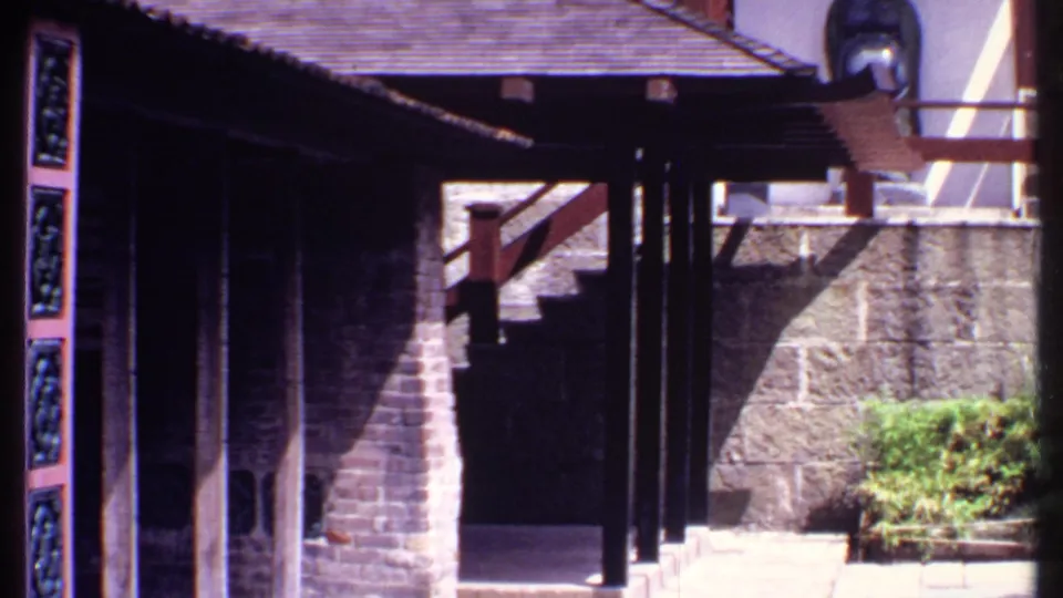 1969 Bahamas resort walkway with dark posts, tiled roof edges, carved blue panels, stone walls, steps, and a sunlit shrub.