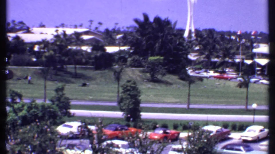 Bahamas resort grounds in 1969 with palms, classic parked cars, a lawn area, low hotel buildings, and a white tower beyond.