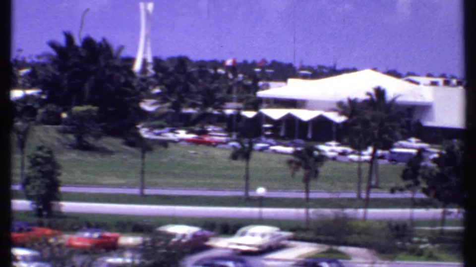 1969 Bahamas hotel exterior with a low white resort building, palms, parked cars, roadway, and landscaped grass.