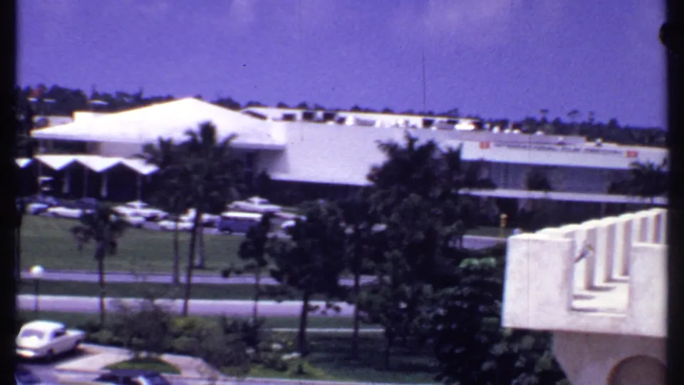1969 Bahamas resort car park view showing classic cars, palm trees, a white hotel building, and part of a nearby balcony or facade.