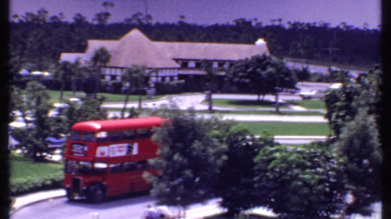 Red double-decker bus passing trees and lawns near low buildings.