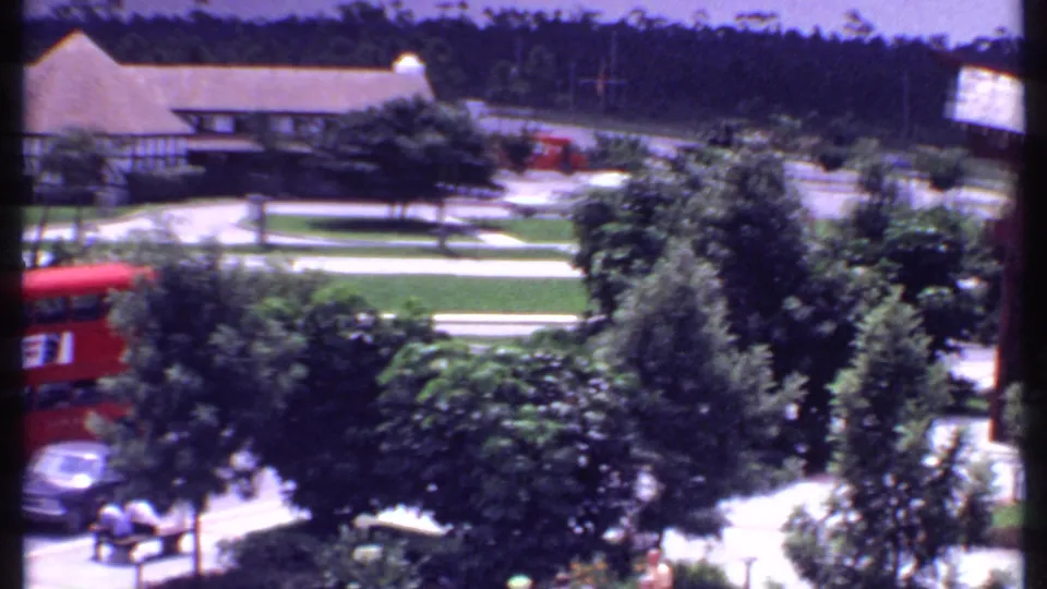 1969 Bahamas 8mm frame showing the red bus partly hidden by trees, with cars and small distant figures near the road.