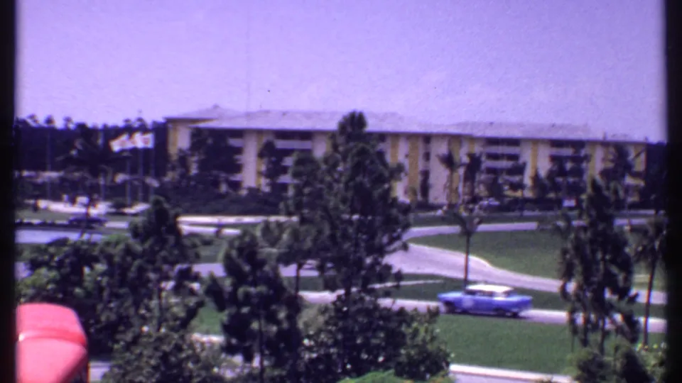1969 Bahamas resort exterior with a long low-rise building, manicured grass, palms, curving paths, and a blue car on the road.