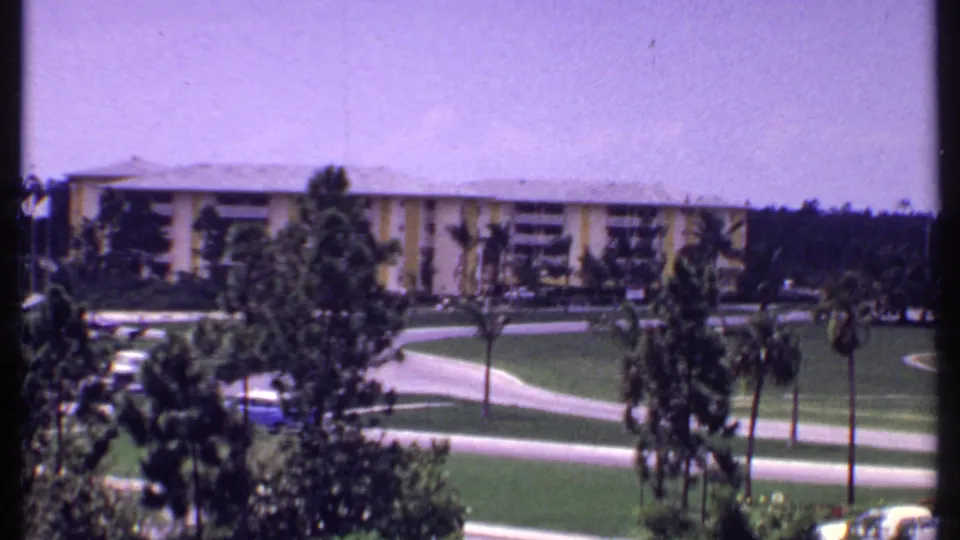 1969 Bahamas home-movie frame showing a resort building behind palm trees, open lawns, winding paths, and cars along the roadway.