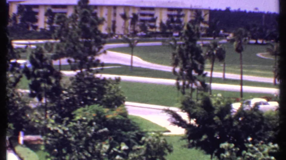 1969 Bahamas resort landscape with palms, clipped lawns, winding walkways, a low-rise building, and light-colored cars at the lower edge.