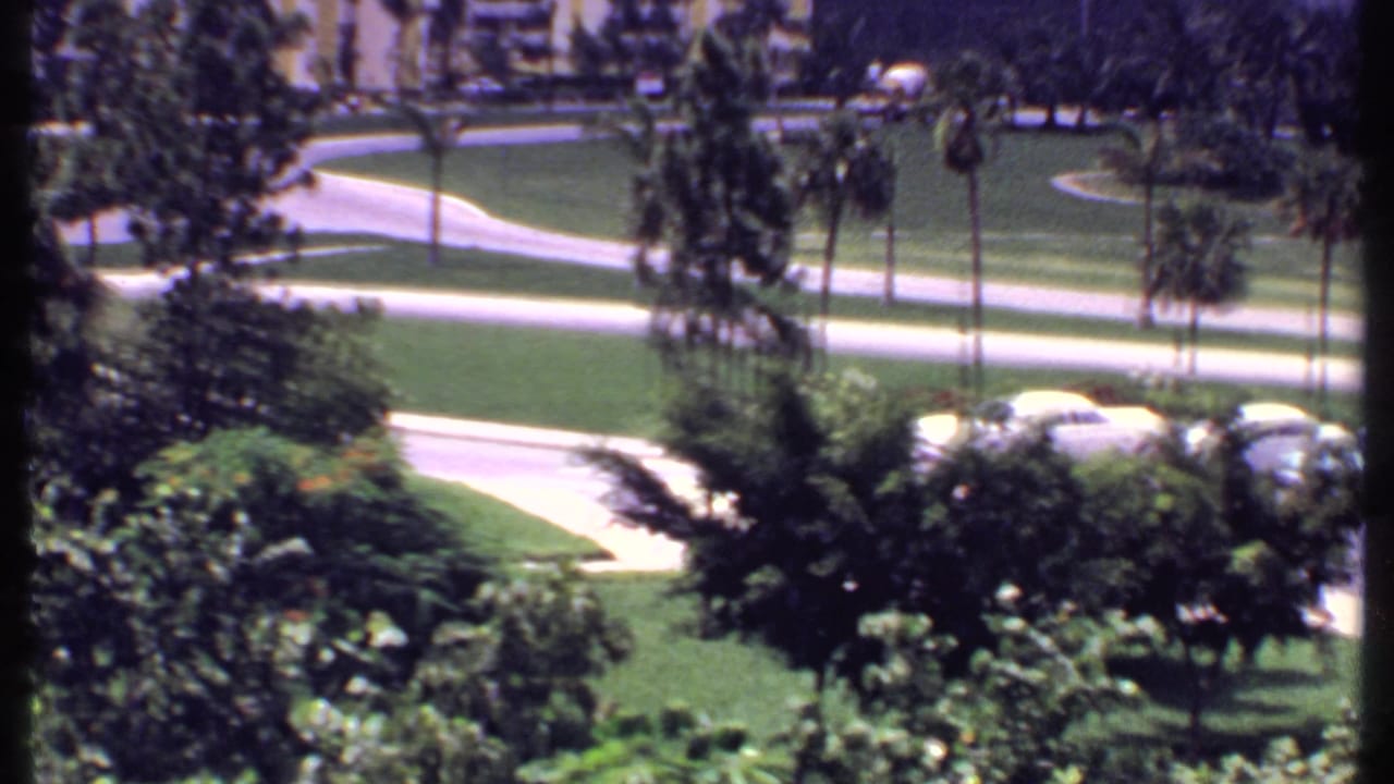 1969 Bahamas home-movie view over landscaped resort grounds with palm trees, paths, roads, and period cars parked nearby.