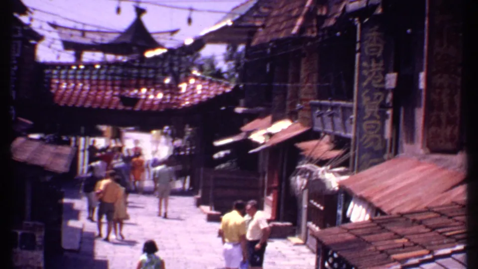 1969 Bahamas market-area scene with thatched structures, hanging signs, and pedestrians walking through a narrow open walkway.