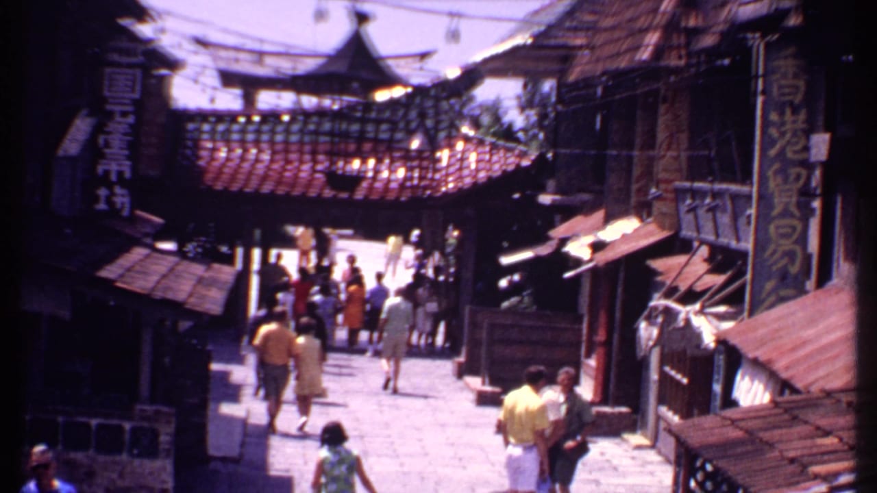 Tourists walk along a stone street beneath Chinese-style tiled roofs and shop signs.