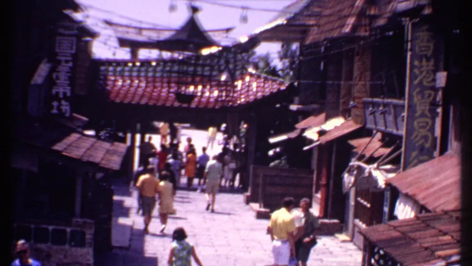 1969 Bahamas home-movie frame showing tourists on a Chinese-themed shopping street with tiled roofs, hanging signs, and shaded storefronts.