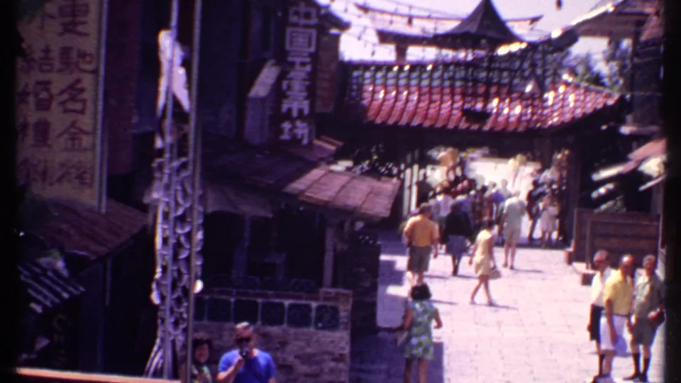 1969 Bahamas travel footage frame with tourists moving past dark shopfronts, vertical signs, tiled rooflines, and a bright stone walkway.