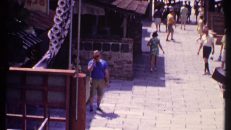 1969 Bahamas frame showing tourists in summer clothing walking through a Chinese-themed street with railings, signs, tiled roofs, and stone paving.