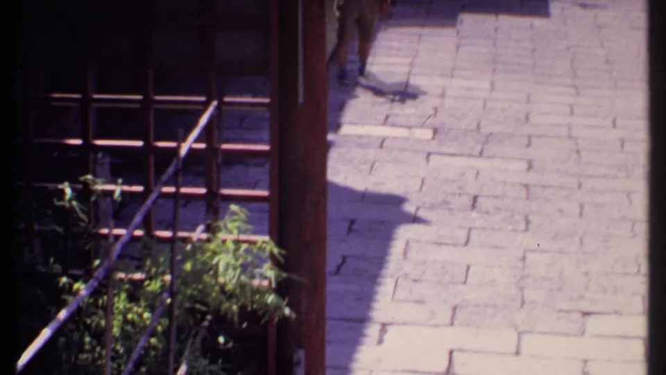 1969 Bahamas home-movie frame with a sunlit stone path, dark wooden structure, potted plants, and tourists farther along the themed street.