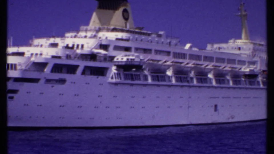 1969 Bahamas-associated 8mm frame showing the side of a white cruise passenger ship with lifeboats, railings, mast, and rows of small windows.