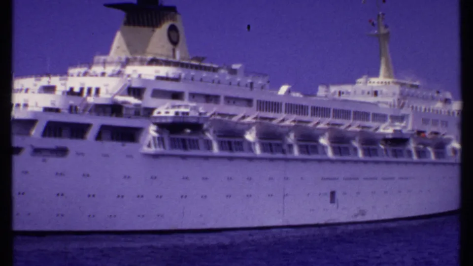 1969 Bahamas 8mm frame showing a large white cruise ship with decks, lifeboats, funnel, and mast above calm ocean water.