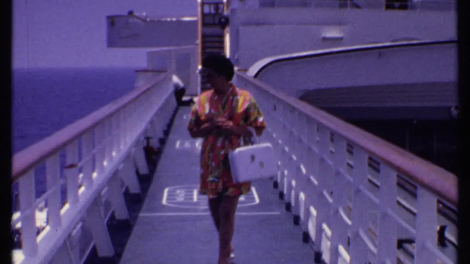 A woman walks toward the camera on a Bahamas cruise ship gangway in 1969, carrying a white handbag beside white railings.