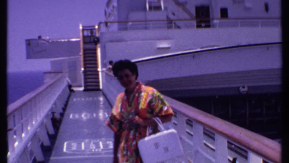 The woman stands nearer the camera at the edge of the frame on the cruise deck, with stairs, railings, and sea visible behind her.