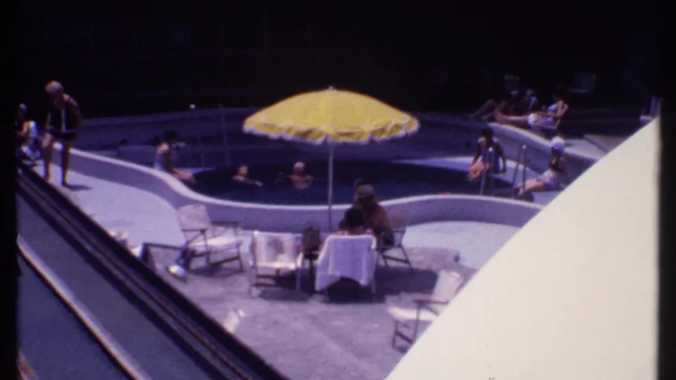 1969 Bahamas cruise frame showing guests seated and swimming around a deck pool beneath a yellow umbrella.