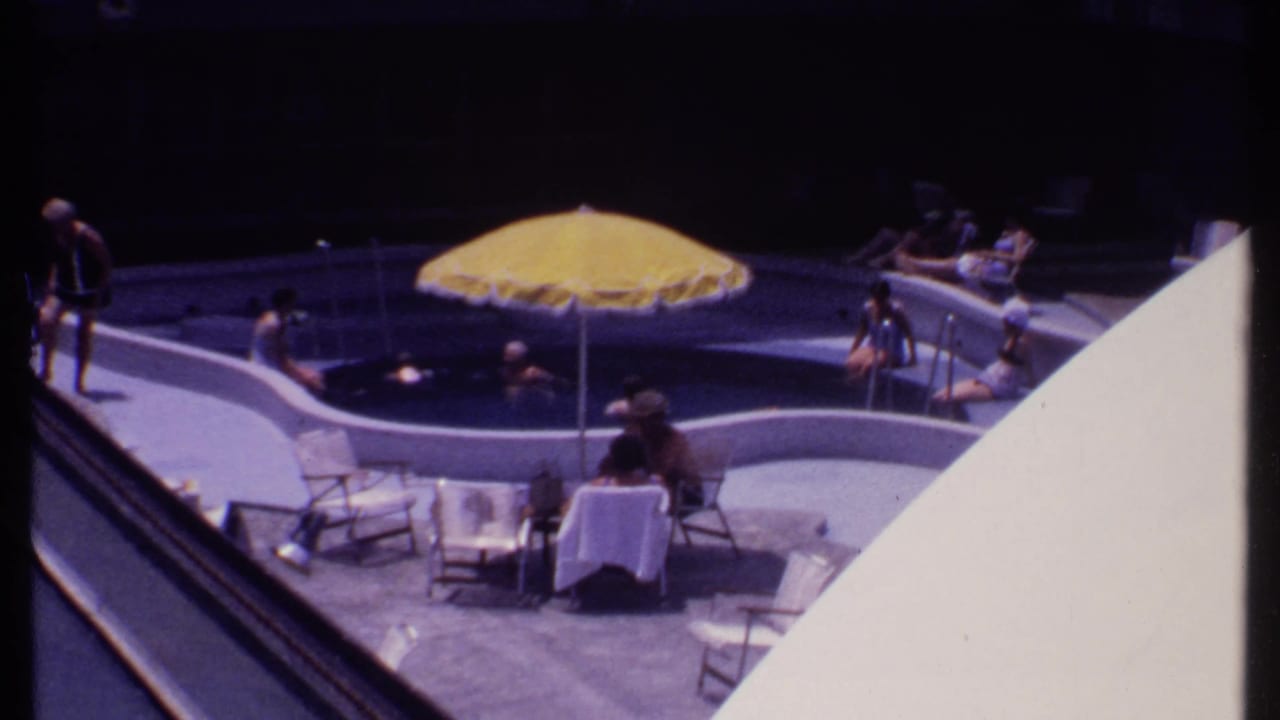 1969 Bahamas home-movie frame showing guests around a curved pool with a yellow umbrella and lounge seating.