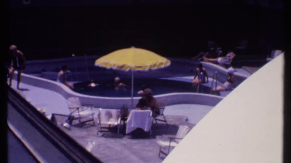 1969 Bahamas home-movie frame showing guests around a curved pool with a yellow umbrella and lounge seating.