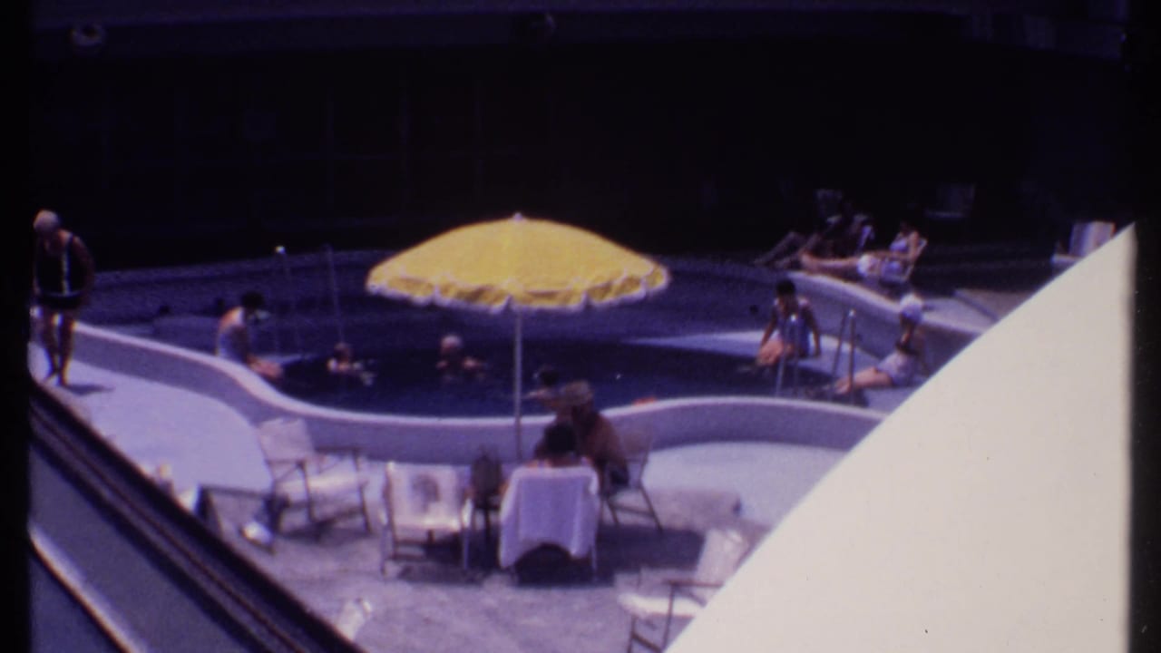 Poolside guests sit and swim near a yellow umbrella and white lounge chairs.