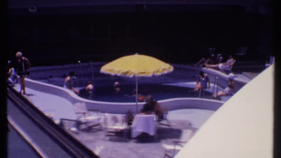 1969 Bahamas vacation frame with a person walking by the curved pool as other guests swim and sit nearby.