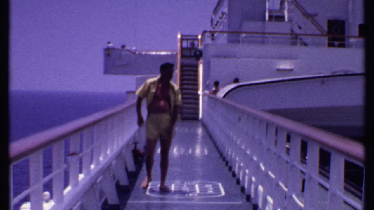 The man walks toward the camera along a white-railed cruise ship deck in the Bahamas, with the sea visible to the left.