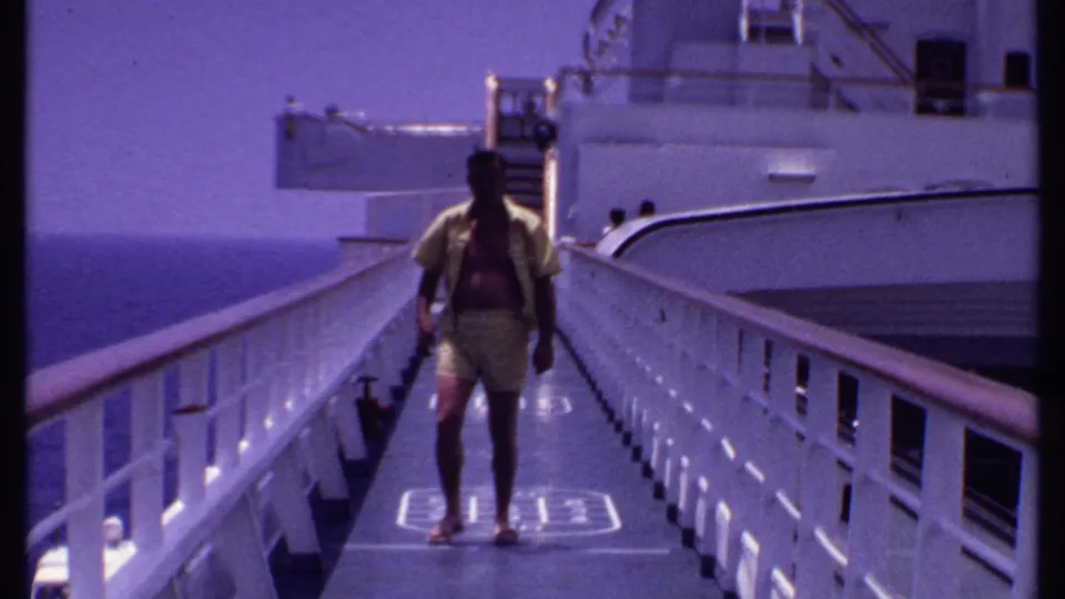 The man continues along the deck toward the camera, passing railings and a shuffleboard marking on the ship deck.