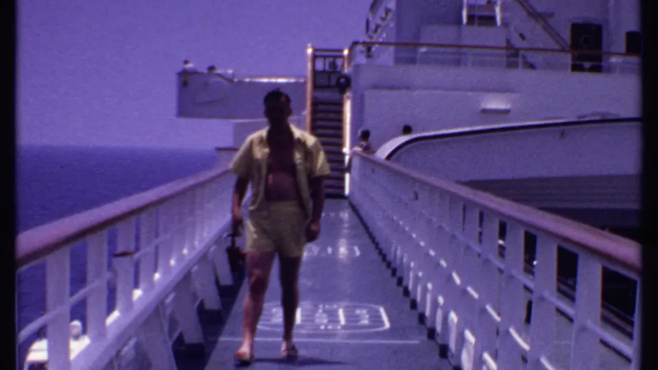At the end of the shot, the man walks past the deck shuffleboard marking with ship railings, stairs, and ocean behind him.