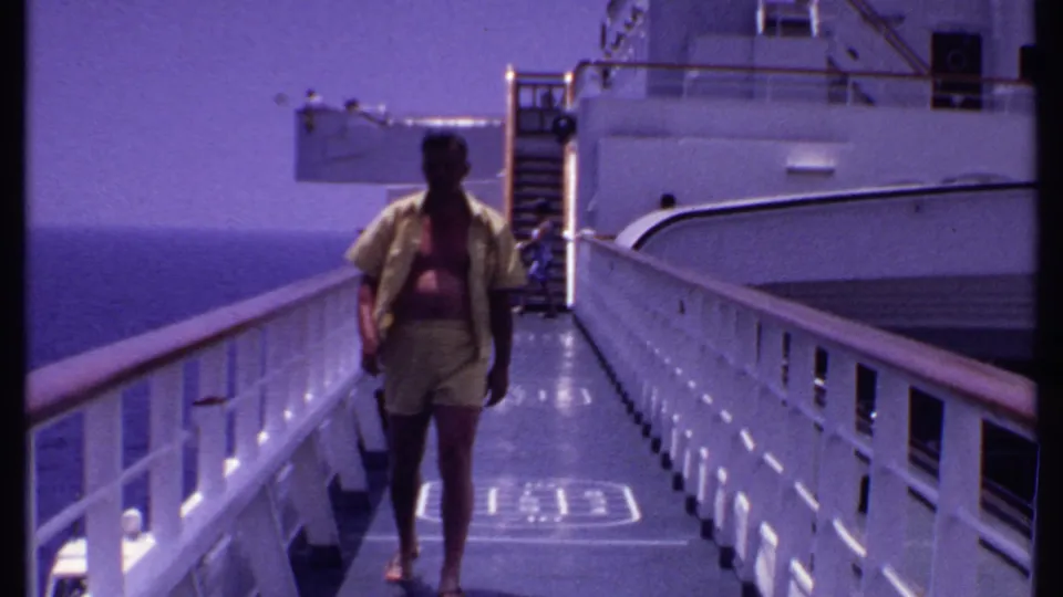 In 1969 Bahamas cruise footage, a man walks toward the camera along a white ship deck with railings and open ocean beyond.