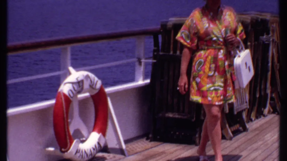 The woman walks along the wooden cruise ship deck with a white handbag, ocean behind the railing, and deck chairs stacked nearby.