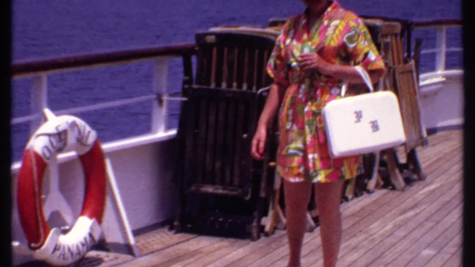 A woman in a colorful 1960s-style dress walks on the cruise ship deck, carrying a white handbag near railings and stacked chairs.