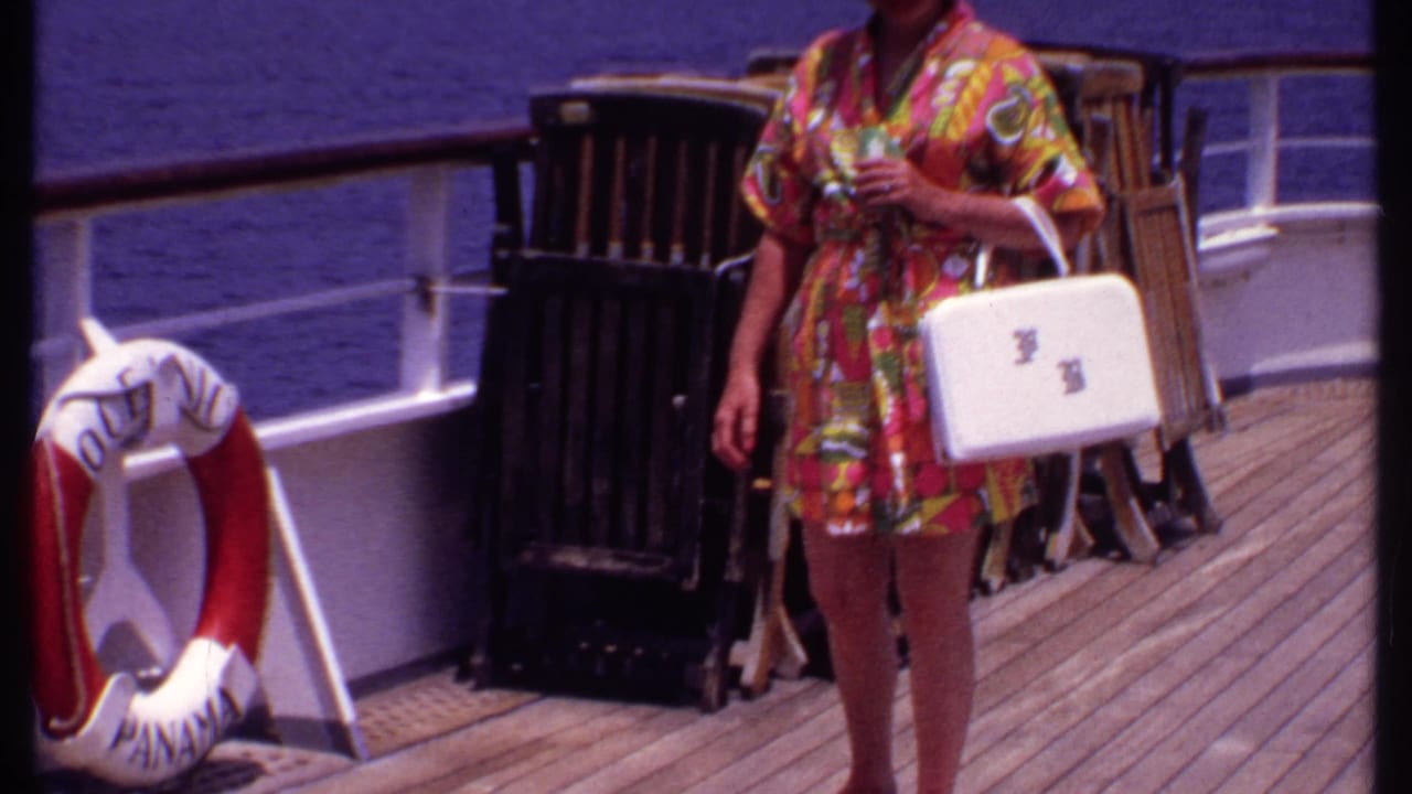 Woman in a bright floral robe stands on a cruise ship deck with a white handbag and drink.