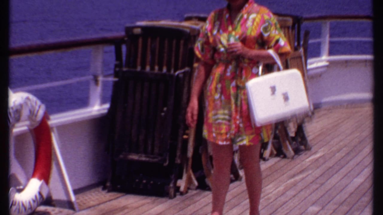 Woman in a floral robe walks along the ship deck past stacked chairs and a life preserver.