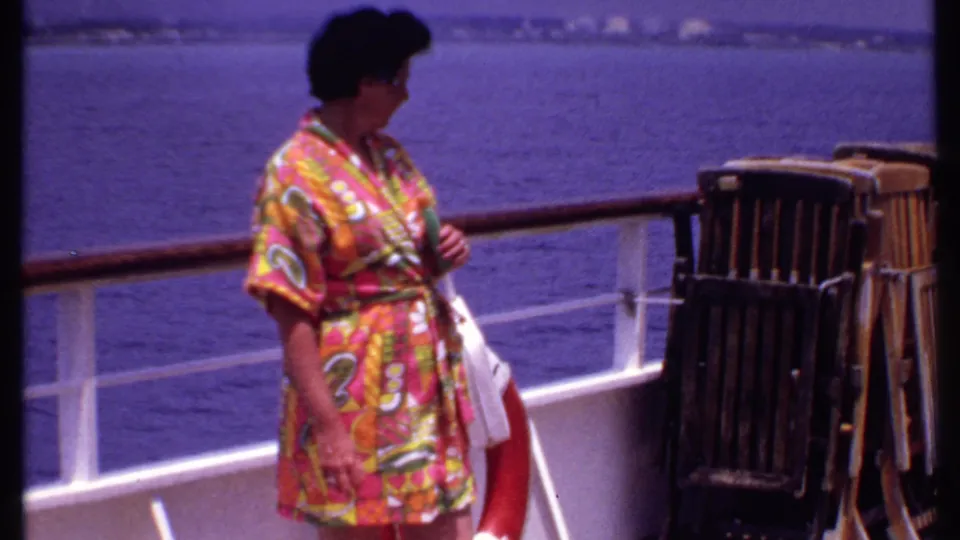 A woman in a colorful floral robe turns on the cruise ship deck, with blue water, white railings, deck chairs, and a life preserver nearby.