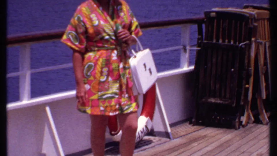 The frame crops close on the cruise passenger’s floral robe, white handbag, legs, ship railing, life preserver, and stacked deck chairs.