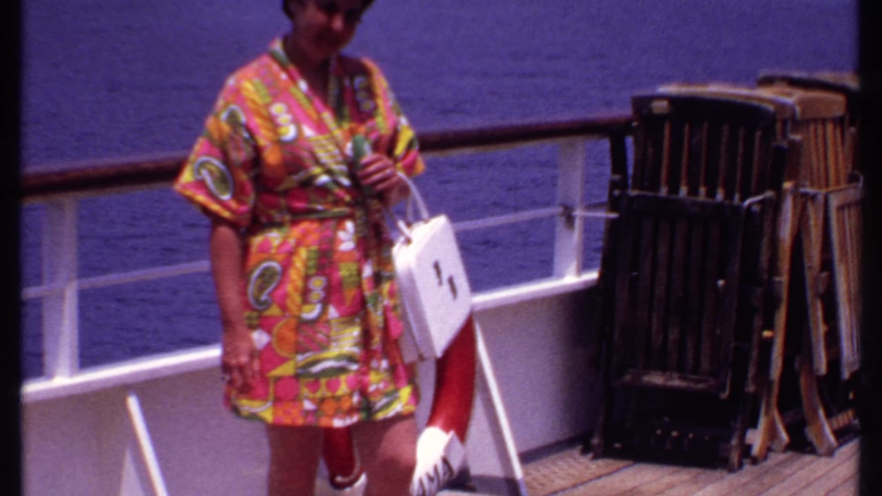 In 1969 Bahamas cruise footage, a woman in a psychedelic robe walks along the deck with a white handbag near railings and stacked chairs.
