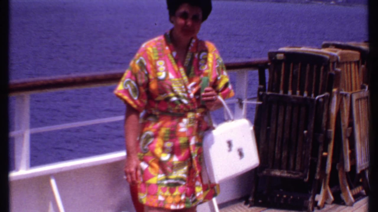 The woman faces the camera on a Bahamas cruise deck, holding a drink and white handbag with the sea visible beyond the railing.