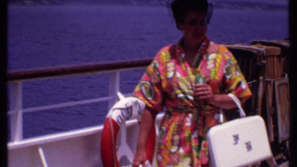 A woman in a colorful robe and sunglasses moves closer to the camera on the cruise deck, carrying a white handbag and a garment.