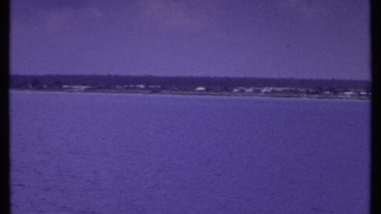 1969 Bahamas home-movie frame with open water in the foreground and a low coastal strip beneath a hazy sky.