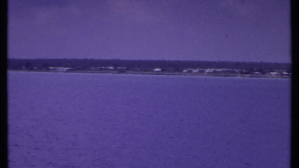1969 Bahamas home-movie frame with open water in the foreground and a low coastal strip beneath a hazy sky.