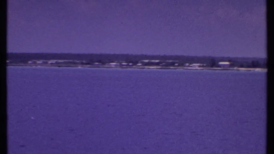 1969 Bahamas travel frame showing calm water, a far tree-lined shore, and pale low structures along the coast.