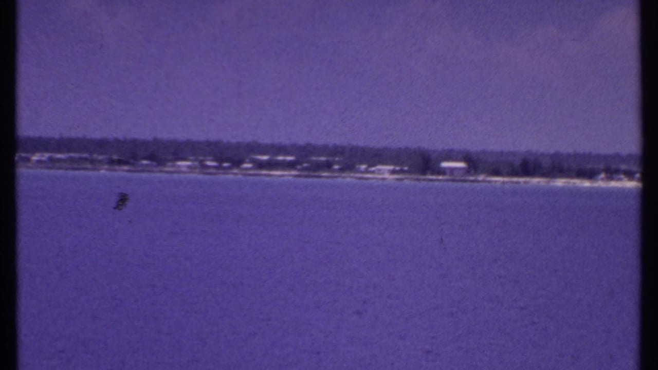 Distant Bahamas shoreline across calm water with beach, trees, and low buildings under a blue sky.