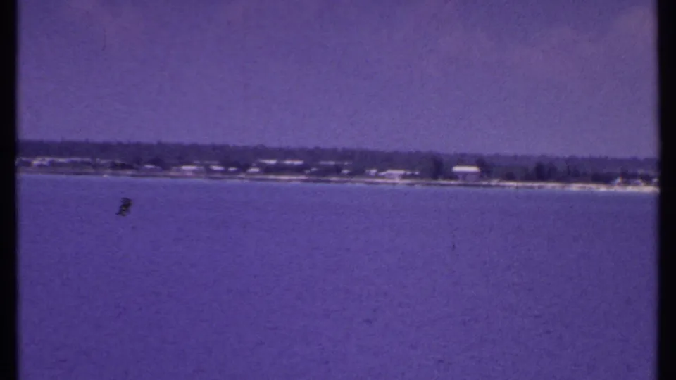 1969 Bahamas 8mm frame showing a distant beach and low waterfront buildings across calm blue water, with a purple vintage color cast.