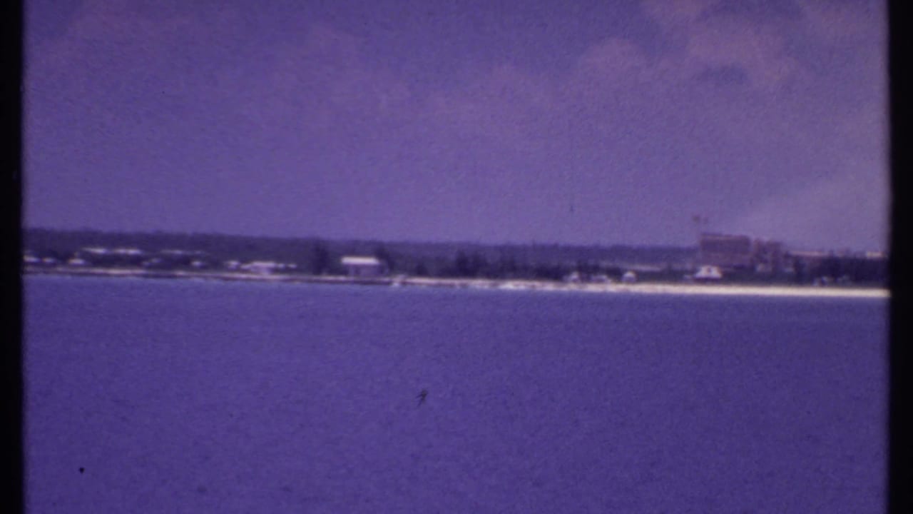 Open water in the foreground with a distant beach, trees, and larger waterfront buildings to the right.