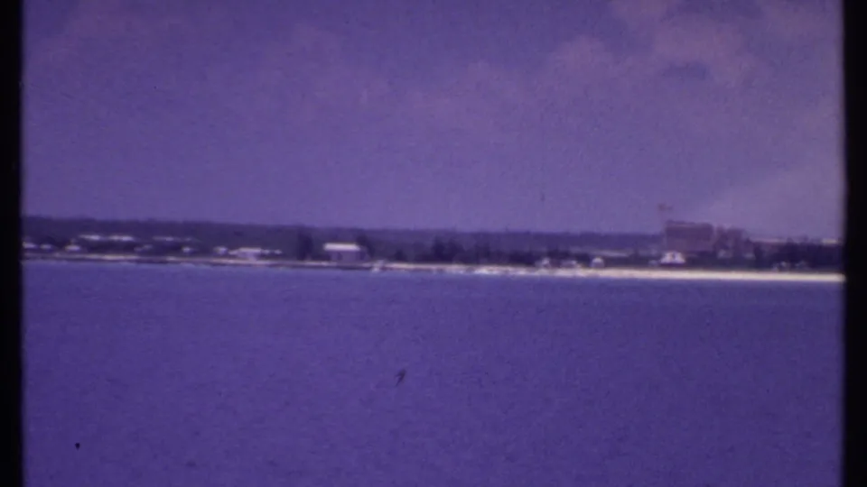 1969 Bahamas shoreline view with calm water, a pale beach strip, trees, and distant low-rise buildings during a rightward pan.